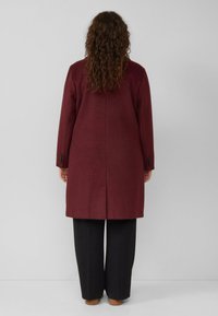 Bordeaux wollen jas met een rechte snit, enkele split en met knopen sluitende manchet, gedragen over zwarte wijde broek. Strak en minimalistisch ontwerp.
