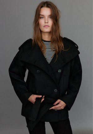 Une femme pose devant un fond de studio gris, vêtue d’un manteau noir ceinturé sur une chemise à rayures noires et blanches.