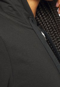 Sweat-shirt à capuche noire avec un tissu texturé, une finition lisse et un cordon de serrage contrastant. Vue rapprochée de la capuche et de la zone des épaules.