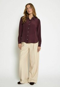 Blusa bordeaux a maniche lunghe con colletto, chiusura frontale con bottoni e tessuto morbido. Abbinata a pantaloni a gamba larga crema e scarpe scure.
