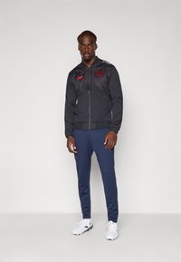 Man som står i en svart zip-up jacka med röda loggor, marinblå byxor och vita fotbollsskor mot en ljus, enfärgad bakgrund.