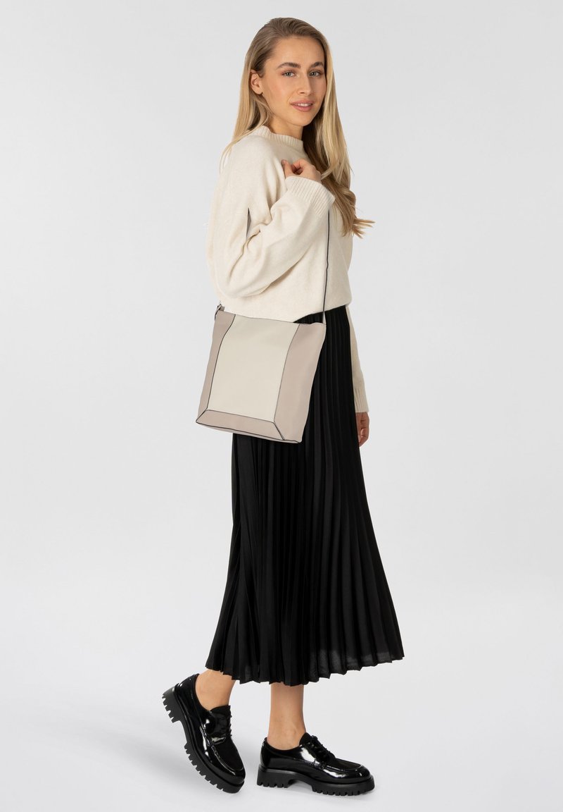 Bolso tote de forma cuadrada en beige y crema, con líneas limpias, una textura suave y una correa desmontable. Llevado con una falda plisada negra.