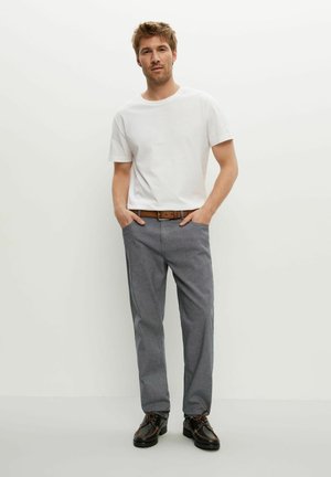 Ein Mann steht mit den Händen in den Taschen, trägt ein weißes Kurzarmhemd, graue Hosen, einen braunen Gürtel und schwarze Schuhe vor einem schlichten hellen Hintergrund.