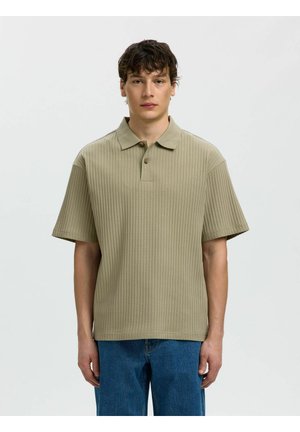 Giovane uomo che guarda in avanti indossa una polo beige a coste con maniche corte e jeans blu su uno sfondo chiaro e semplice.