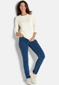 Lächelnde Frau steht da, trägt einen strukturierten cremefarbenen Pullover, blaue enge Jeans und weiße Sneakers, mit einer Hand in der Tasche.