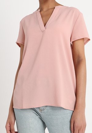 Persoon met een lichtroze V-hals blouse met korte mouwen en lichtblauwe spijkerbroek, staand tegen een effen witte achtergrond.