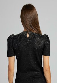 Czarny top z krótkim rękawem, charakteryzujący się detalem w formie otworu w plecach oraz rozproszonymi metalicznymi akcentami, o fakturowanym wykończeniu i bufiastym designie ramion.