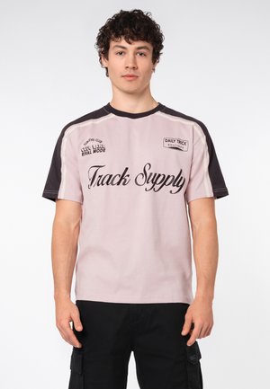 Junger Mann mit lockigem Haar, der ein hellrosa-schwarzes T-Shirt mit dem Schriftzug „Track Supply“ und schwarze Cargoshorts trägt, steht vor weißem Hintergrund.