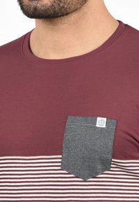 Burgundy t-shirt with a round neck, featuring a grey denim chest pocket and horizontal white and burgundy stripes at the bottom.