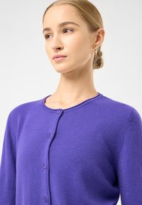 Ung kvinde med blond hår i en lav knold, iført guldøreringe og en lilla knap cardigan, som kigger lidt til siden.