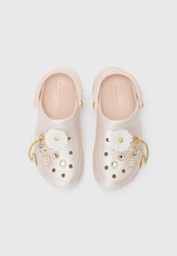 Crocs rose clair à finition brillante, ornés d'embellissements décoratifs floraux et de bijoux, avec une bride arrière ajustable et des trous de ventilation.