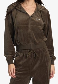 Hoodie marrone con zip in morbido e peloso tessuto, con tasca a marsupio, cappuccio con cordino e dettagli a coste sui polsini. Logo sul lato del petto.