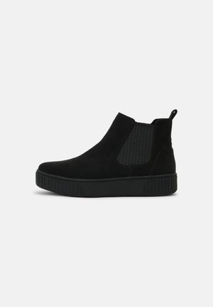 Sort mokkasin ankelstøvle med tyk gummisål, elastisk sidepanel og trækflap bagpå, vist mod en ensfarvet hvid baggrund.