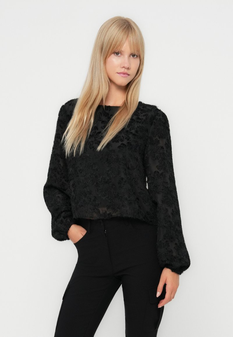 Czarna bluzka z długim rękawem, z teksturowanym kwiatowym wzorem, przezroczystym materiałem i luźnym krojem. W zestawie z dopasowanymi czarnymi spodniami.