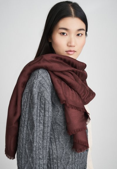 Cachecol borgonha com bordas desfiadas colocado sobre um suéter cinza em malha trançada. O cachecol apresenta um design delicado e padronizado.