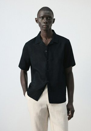 Homme portant une chemise noire à manches courtes boutonnée et un pantalon beige clair, debout avec une main dans la poche devant un fond uni.