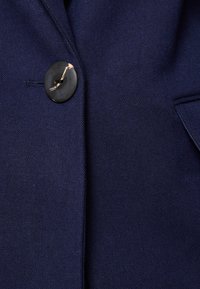 Marinblått tyg med en texturerad väv har en enda svart knapp med en gyllene accent. Tyget har en sl ät yta och en strukturerad design.