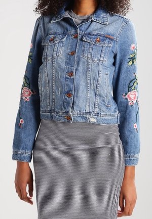 Vrouw draagt een blauwe spijkerjas met bloemrijke borduursels op de mouwen over een zwart-wit gestreepte jurk met horizontale strepen.