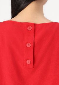 Červený oděv s širokým výstřihem a třemi červenými knoflíky vzadu. Tkanina vypadá hladce a lehce. Jednoduchý design bez vzorů.
