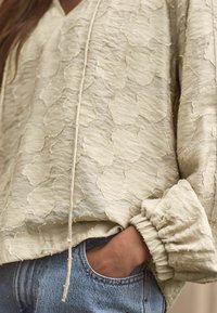 Helles beige Bluse mit Struktur und subtilen Blumenmuster, Kordelzug-Detail und gerafften Ärmeln. Kombiniert mit blauen Jeans.