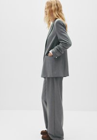 Sacou tailored gri și pantaloni evazați dintr-un material moale, texturat. Prezintă buzunare adânci și este accesorizat cu pantofi de tip mocasin de culoare maronie.