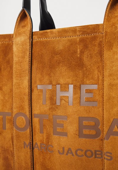 Brązowa skórzana torba na ramię z czarnymi skórzanymi uchwytami. Na przodzie znajduje się duży, wypukły napis "TORBA NA RAMIĘ MARC JACOBS".