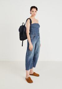 Blue denim jumpsuit with cropped wide legs, featuring a strapless design and front seams. Paired with a black backpack and brown clogs.