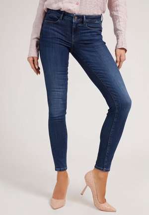 Kobieta nosząca ciemnoniebieskie dopasowane dżinsy w połączeniu z jasnoróżowymi wzorzystymi butami na wysokim obcasie i blado różową bluzką z długim rękawem.