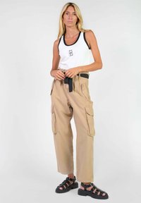 Witte tanktop met zwarte rand en logo, gecombineerd met beige cargo broek met zijzakken en een zwarte riem. Zwarte sandalen met bandjes.