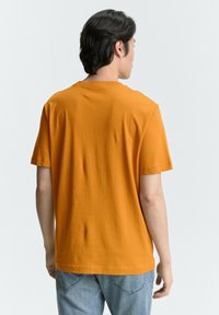 Camiseta de manga corta y cuello redondo en algodón de color naranja brillante. Textura suave, corte regular, mostrada desde la parte de atrás con pantalones de mezclilla claros.