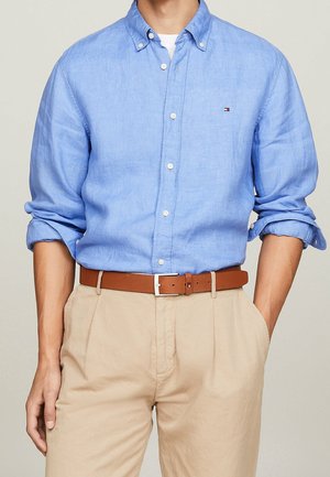 Homme portant une chemise bleu clair à boutons avec les manches retroussées, pantalon beige et ceinture en cuir marron, debout avec une main dans la poche.