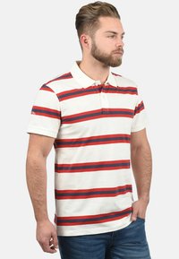 Korte mouw polo shirt in crème met horizontale rode en marineblauwe strepen. Voorzien van een kraag en stofstructuur die kenmerkend is voor katoen.