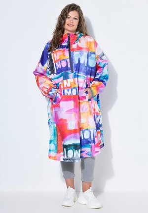 Kvinde iført en farverig oversized jakke med dristige tekstmønstre, grå cropped bukser og hvide sneakers, stående mod en hvid baggrund.