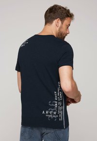 Camiseta azul marino de manga corta, con texto y gráficos blancos a lo largo del costado. La tela parece suave y tiene un ajuste casual.