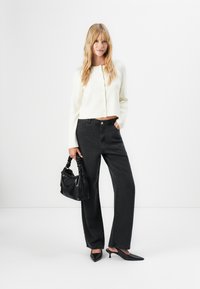 Blonde vrouw in een witte gesloten cardigan, zwarte high-waisted wijde broek, zwarte hakken, met een zwarte handtas.