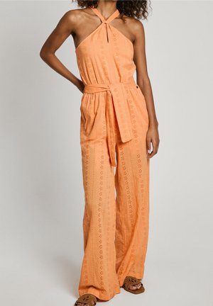 Femme portant une combinaison sans manches orange à col halter avec un motif œillet et une taille nouée, associée à des sandales marron à bouts ouverts.