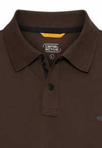 Bruine Camel Active polo met zwarte knopen, zwarte binnenkraagafwerking en maatlabel "L" zichtbaar binnenin de halslijn.