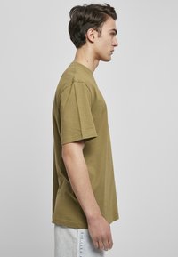 Tricou din bumbac de culoare verde măsliniu, cu mâneci scurte și decolteu rotund, expus în profil lateral pe un fundal gri.