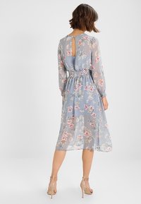 Žena v světle modrých květovaných midi šatech s průhlednými dlouhými rukávy, s nařaseným pasem a vykrojeným zádovým detailem, doplněná o tělové lodičky.