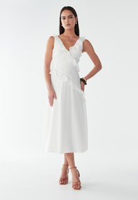 Weißes Midikleid mit rüschiertem Ausschnitt und Saum, aus leichtem Stoff gefertigt. Kombiniert mit braunen Riemchen-Heels und minimalem Schmuck.