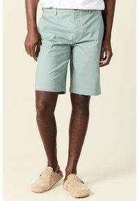 Pantalones cortos verdes claros hechos de tejido texturizado, con un corte recto, bolsillos laterales y parte delantera plana. Usados con zapatos beige tipo slip-on.