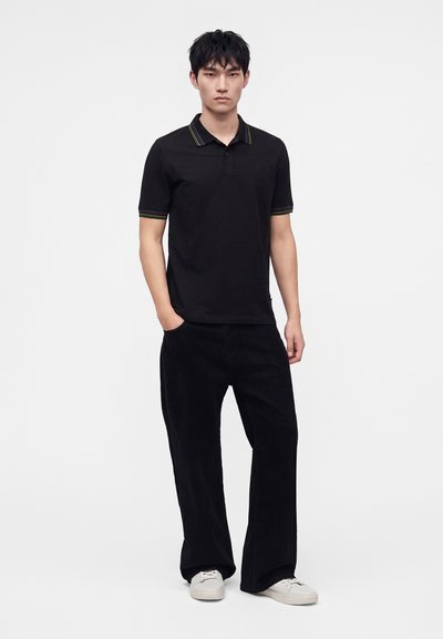 Jeune homme debout, une main dans la poche, portant un polo noir avec des bords rayés, un pantalon noir ample et des baskets blanches sur fond blanc.