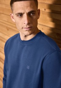 Blå sweatshirt med rund halsringning och långa ärmar, med en diskret logga på bröstet. Tyget ser mjukt ut mot en träbakgrund.