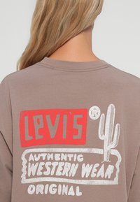 Smeđa oversize majica s grafikom na leđima koja prikazuje "Levi's" u crvenoj boji, "Authentic Western Wear" u bijeloj boji i ilustraciju kaktusa.