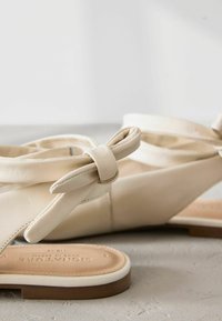 Beige leather sandals with a knot detail, open toe design, and a textured sole featuring a light beige footbed and brown outer sole.