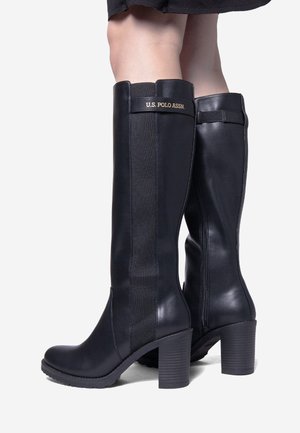 Mustat polvenkorkuiset korkokengät joustavilla sivupaneeleilla, tukevilla koroilla ja "U.S. Polo Assn." -logolla, yhdistettynä mustaan hameeseen.