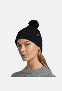 Gorro de punto acanalado negro con un pompón en la parte superior y un parche circular de logo. Lo lleva puesto un modelo, mostrando su ajuste ceñido y textura.