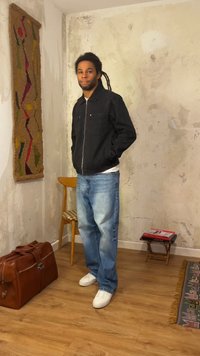 Νέος άνδρας με ντρέντλοκς στέκεται σε ένα δωμάτιο με φθαρμένους τοίχους, φοράει μαύρο μπουφάν, μπλε τζιν και λευκά αθλητικά παπούτσια.