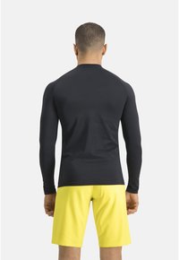 Schwarzes, langärmliges Sportshirt aus glattem Material, kombiniert mit gelben Shorts. Das Shirt hat ein tailliertes Design und Raglanärmel.