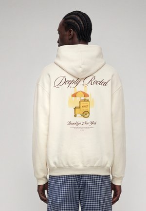 Homme portant un sweat à capuche crème avec le texte "Deeply Rooted" et un graphisme de charrette à hot-dogs jaune, associé à un pantalon bleu à carreaux, vu de dos.